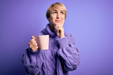 Young blonde woman with short hair wearing winter sweater drinking a cup of hot coffee serious face thinking about question, very confused idea