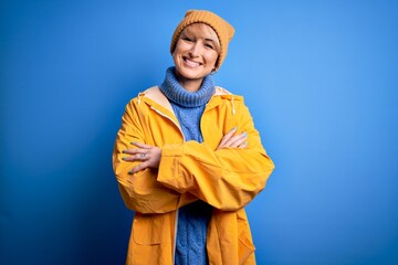 Young blonde woman with short hair wearing rain coat for rainy weather over blue background happy...