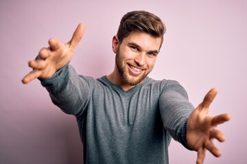 Young handsome blond man with beard and blue eyes wearing casual sweater looking at the camera smiling with open arms for hug. Cheerful expression embracing happiness.