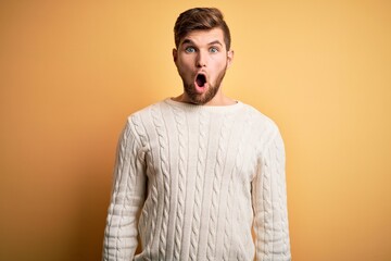 Young blond man with beard and blue eyes wearing white sweater over yellow background afraid and shocked with surprise and amazed expression, fear and excited face.