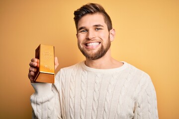 Young blond man with beard and blue eyes holding gold ingot over isolated yellow background with a...