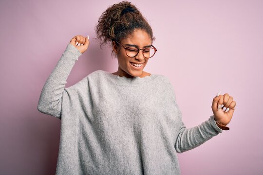 Young beautiful african american girl wearing sweater and glasses over pink background Dancing happy and cheerful, smiling moving casual and confident listening to music