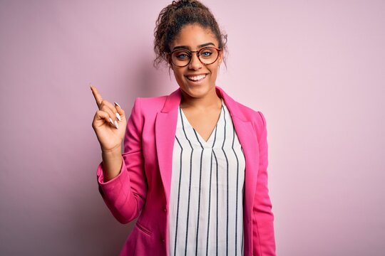 Beautiful african american businesswoman wearing jacket and glasses over pink background with a big smile on face, pointing with hand finger to the side looking at the camera.