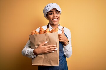 Young african american baker girl wearing apron holding bag with french croissants happy with big smile doing ok sign, thumb up with fingers, excellent sign