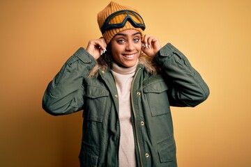 Young african american afro skier girl wearing snow sportswear and ski goggles Smiling pulling ears with fingers, funny gesture. Audition problem