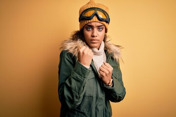 Young african american afro skier girl wearing snow sportswear and ski goggles Ready to fight with fist defense gesture, angry and upset face, afraid of problem