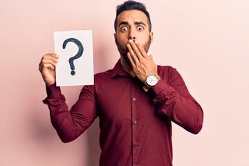 Young hispanic man holding question mark covering mouth with hand, shocked and afraid for mistake. surprised expression