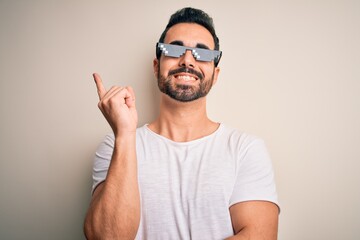 Fototapeta premium Young handsome man with beard wearing funny thug life sunglasses over white background with a big smile on face, pointing with hand and finger to the side looking at the camera.