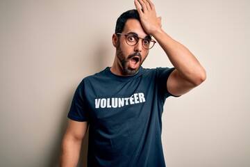 Handsome man with beard wearing t-shirt with volunteer message over white background surprised with hand on head for mistake, remember error. Forgot, bad memory concept.