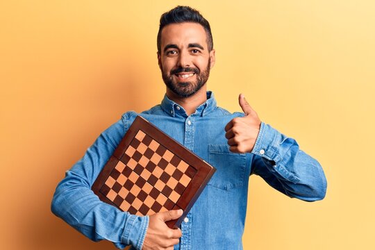 Young hispanic man holding chess smiling happy and positive, thumb up doing excellent and approval sign