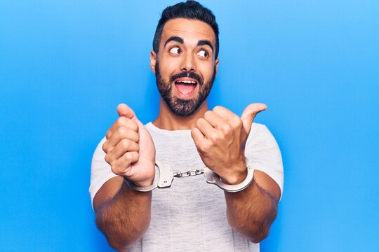 Young hispanic man wearing prisoner handcuffs pointing thumb up to the side smiling happy with open mouth