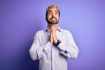 Young handsome man with beard wearing golden crown of king over purple background begging and praying with hands together with hope expression on face very emotional and worried. Begging.