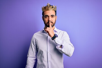 Young handsome man with beard wearing golden crown of king over purple background asking to be quiet with finger on lips. Silence and secret concept.