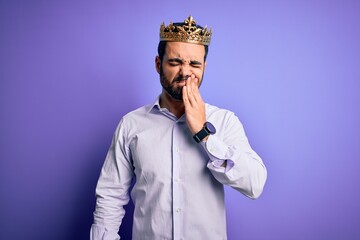 Young handsome man with beard wearing golden crown of king over purple background touching mouth with hand with painful expression because of toothache or dental illness on teeth. Dentist