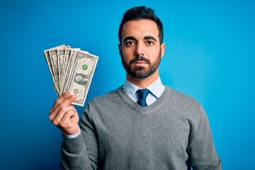 Young handsome man with beard holding bunch of dollars banknotes over blue background with a confident expression on smart face thinking serious