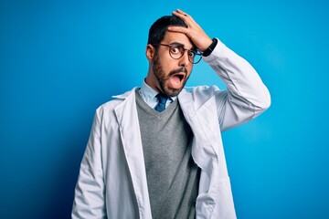 Young handsome doctor man with beard wearing coat and glasses over blue background surprised with...