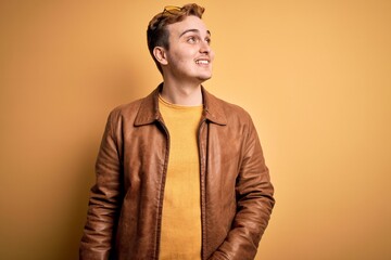 Young handsome redhead man wearing casual leather jacket over isolated yellow background looking away to side with smile on face, natural expression. Laughing confident.