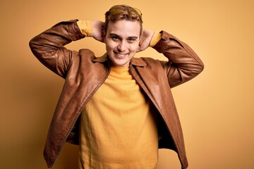 Young handsome redhead man wearing casual leather jacket over isolated yellow background relaxing and stretching, arms and hands behind head and neck smiling happy