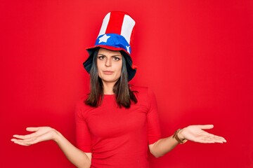 Young beautiful brunette woman wearing united states hat celebrating independence day clueless and confused with open arms, no idea concept.