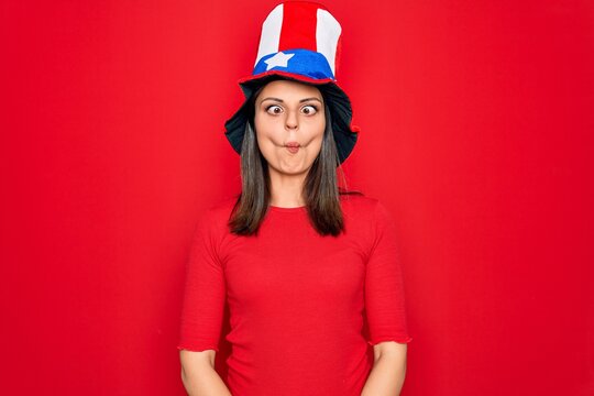 Young beautiful brunette woman wearing united states hat celebrating independence day making fish face with lips, crazy and comical gesture. Funny expression.