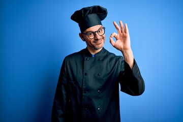 Young handsome chef man wearing cooker uniform and hat over isolated blue background smiling positive doing ok sign with hand and fingers. Successful expression.