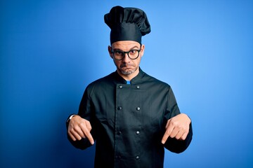 Young handsome chef man wearing cooker uniform and hat over isolated blue background Pointing down looking sad and upset, indicating direction with fingers, unhappy and depressed.