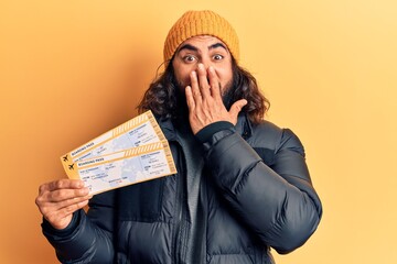 Young arab man holding boarding pass covering mouth with hand, shocked and afraid for mistake....