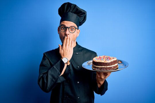 Young handsome baker man wearing cooker uniform and hat holding tray with cake cover mouth with hand shocked with shame for mistake, expression of fear, scared in silence, secret concept