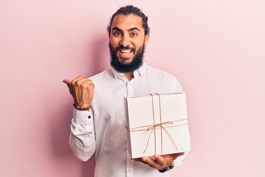 Young arab man holding gift pointing thumb up to the side smiling happy with open mouth