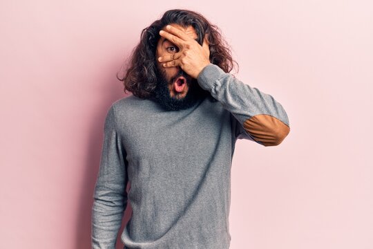 Young arab man wearing casual clothes peeking in shock covering face and eyes with hand, looking through fingers afraid