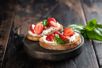 Two bruschettas with strawberries, cream cheese and basil on a round wooden board on an old wooden background