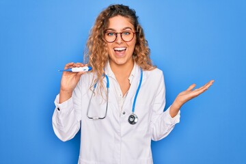 Young beautiful blonde doctor woman with blue eyes wearing stethoscope holding thermometer very happy and excited, winner expression celebrating victory screaming with big smile and raised hands