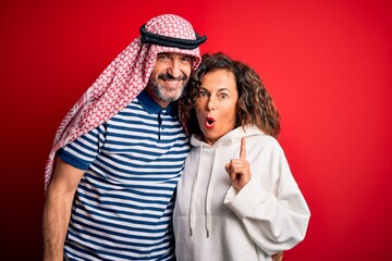 Middle age beautiful couple wearing arab headscarf and sweatshirt smiling happy and confident. Standing with smile on face hugging over isolated red background