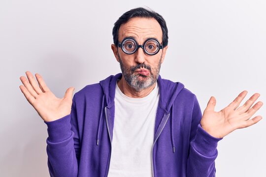 Middle age handsome freaky man wearing dumb glasses over isolated white background clueless and confused with open arms, no idea and doubtful face.