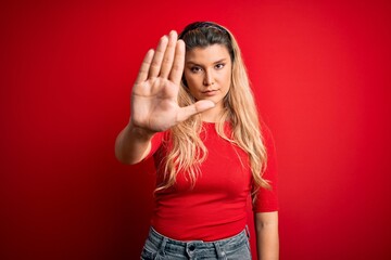Young beautiful blonde woman wearing casual t-shirt standing over isolated red background doing stop sing with palm of the hand. Warning expression with negative and serious gesture on the face.