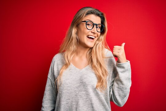 Young beautiful blonde woman wearing sweater and glasses over isolated red background smiling with happy face looking and pointing to the side with thumb up.