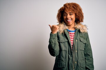 Young beautiful African American woman with curly hair wearing green casual winter coat smiling with happy face looking and pointing to the side with thumb up.