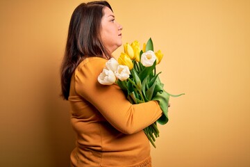 Beautiful plus size woman holding romantic bouquet of natural tulips flowers over yellow background looking to side, relax profile pose with natural face and confident smile.