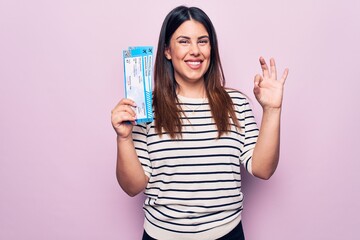 Young beautiful tourist woman holding airline boarding pass over isolated pink background doing ok sign with fingers, smiling friendly gesturing excellent symbol
