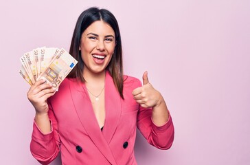 Young beautiful businesswoman holding euros banknotes over isolated pink background smiling happy and positive, thumb up doing excellent and approval sign