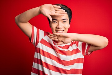 Fototapeta premium Young handsome chinese man wearing casual striped t-shirt standing over red background Smiling cheerful playing peek a boo with hands showing face. Surprised and exited