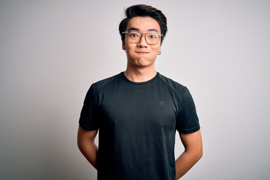 Young handsome chinese man wearing black t-shirt and glasses over white background puffing cheeks with funny face. Mouth inflated with air, crazy expression.