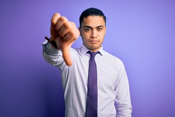 Young brazilian businessman wearing elegant tie standing over isolated purple background looking unhappy and angry showing rejection and negative with thumbs down gesture. Bad expression.