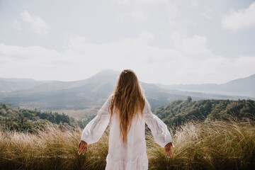 A woman in a white dress looks at the volcano with her arms outstretched. Rear view. The Agung...