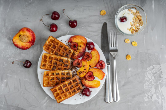 White plate full of belgian wafers and mix slices of cherries and peaches together with yogurt and cutlery are laying on concrete surface