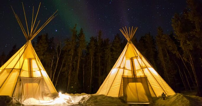 Time lapse shot of illuminated teepees on snow by trees against sky at night - Northwest Territories, Canada