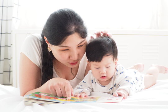 Asian mother storytelling and teaching her cute newborn baby with cartoon picture book in the bedroom at home 