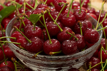 red cherries in a transparent glass bowl on a wooden background, with green leaves of a cherry tree. Summer berries. Yummy.