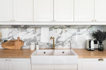 Modern classic kitchen interior with kitchen appliances and white ceramic sink with gold mirror faucet on wood top with marble wall in the background / kitchen interior