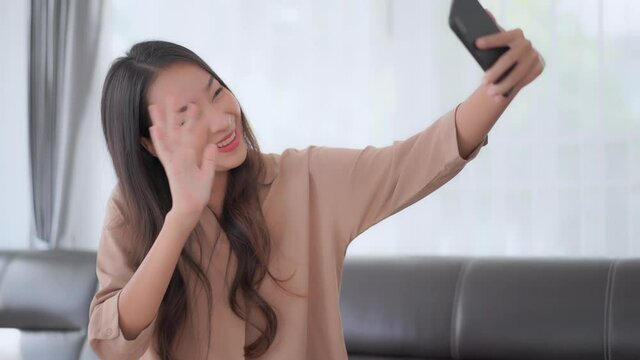 Closeup shot of adult Asian woman having a video call on the phone indoors in the living room, sitting on sofa, showing palm and Ok gesture 